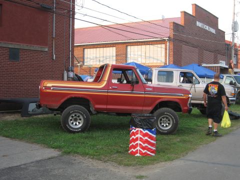 1984 Ford Bronco - Red Ford Bronco with racing stripes parked on grass next to a building. Another truck is parked behind it.