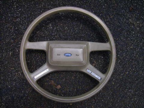1982 Ford LTD Crown Victoria - A tan four-spoke Ford steering wheel lying face-up on asphalt, its rectangular center horn pad carrying the blue Ford oval flanked by horn…