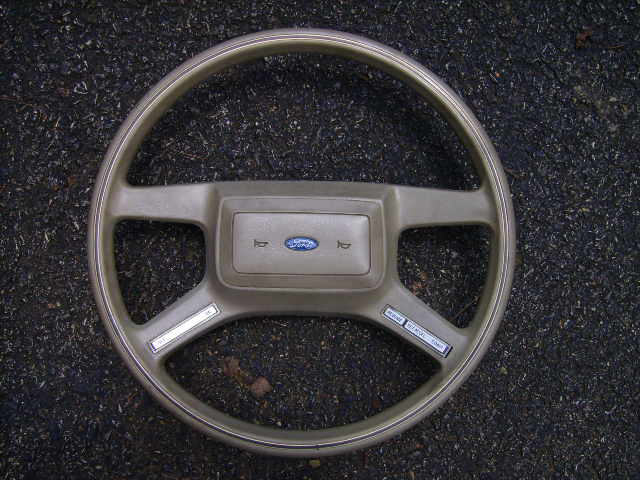 Tan four-spoke Ford steering wheel with blue Ford oval horn pad, laid on asphalt