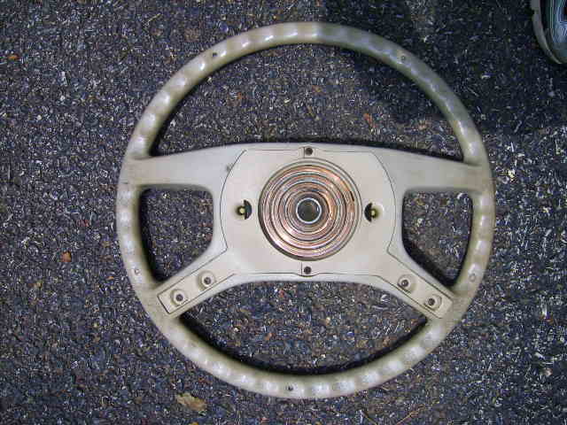 Removed tan four-spoke steering wheel on asphalt with horn pad off, exposing contact ring