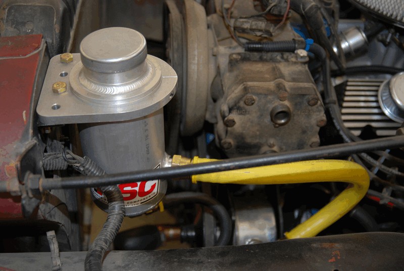 Engine bay with a silver component mounted on a bracket, yellow hose, and black wiring