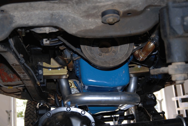 Underside view of a vehicle's engine, showing the oil pan, exhaust pipes, and drive belt