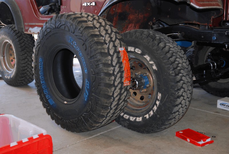Two large off-road tires, one mounted on a wheel, in front of a red Jeep CJ