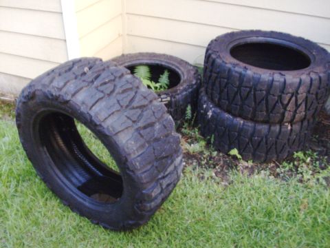 1995 Ford Bronco - A set of four unmounted mud-terrain tires with chunky lug tread stacked and leaned against the beige siding of a house on a grass lawn. Fer…