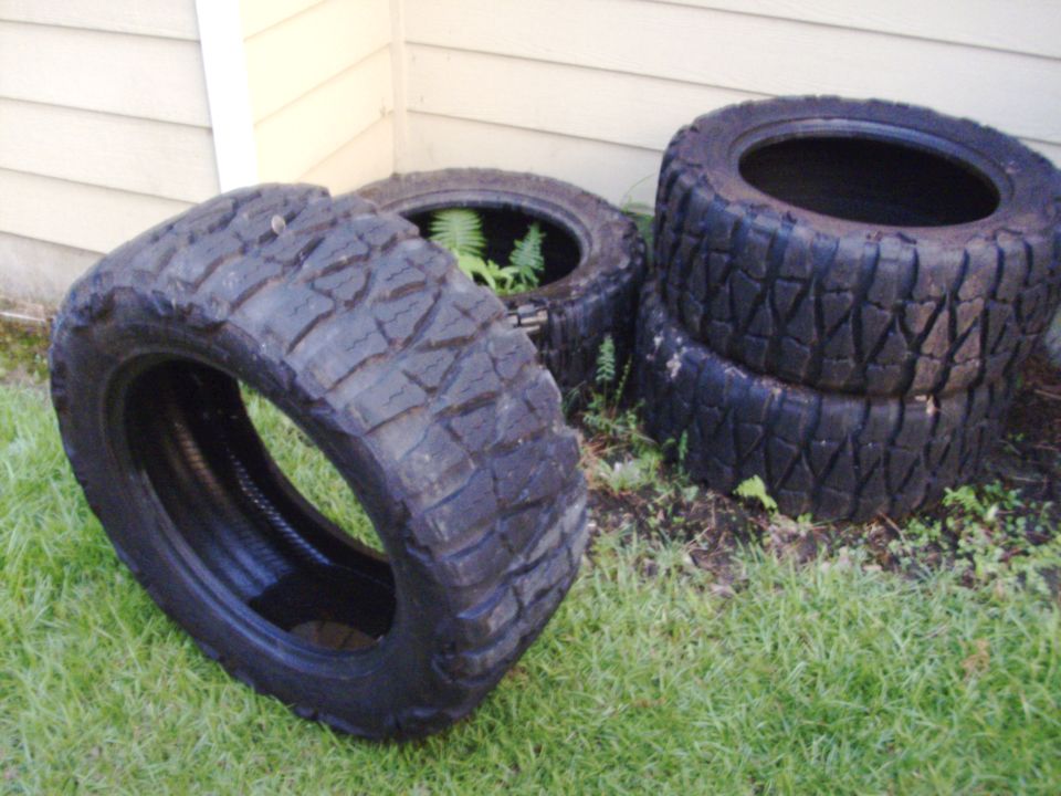 Four used unmounted mud-terrain tires stacked in grass against beige house siding