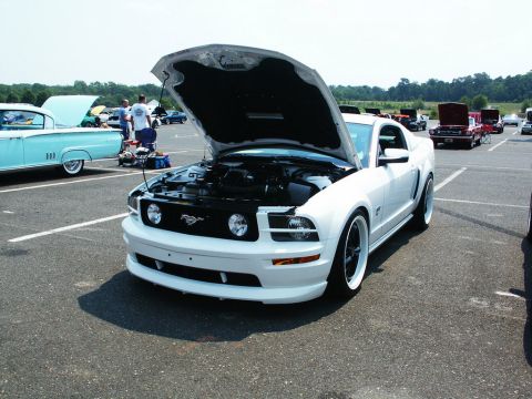 2001 Mercury Grand Marquis - A white Ford Mustang is displayed with its hood open at an outdoor car show, with other vehicles visible in the background.