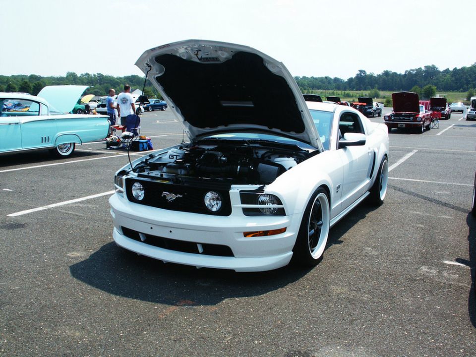White Ford Mustang with hood open, parked at a car show. Another classic car is to the left