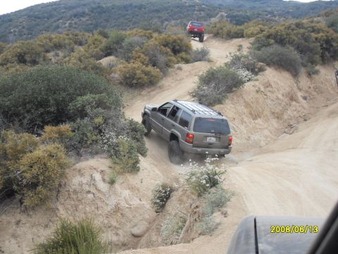 1990 Ford Bronco - A gray Jeep Grand Cherokee navigates a steep, dusty off-road trail, with another vehicle visible further up the path.
