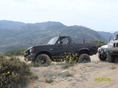 1990 Ford Bronco - A black Ford Bronco is parked on a dirt road next to another vehicle, with mountains in the background.