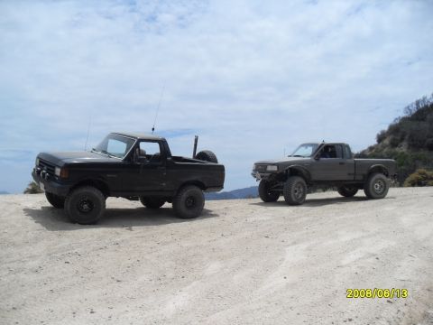 1990 Ford Bronco - Two off-road vehicles, a black 1990 Ford Bronco and a grey pickup, are parked on a dusty trail with a scenic background.