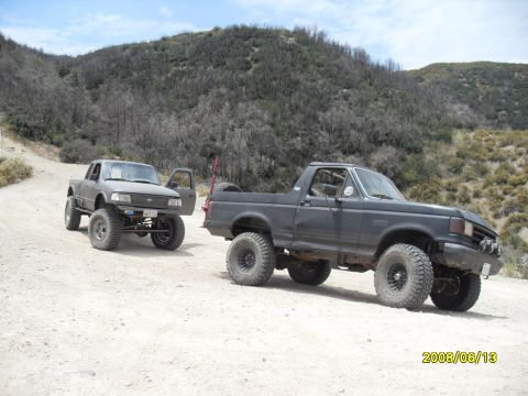 1990 Ford Bronco - Two modified trucks are parked on a dusty trail with hills in the background.