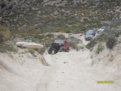 1990 Ford Bronco - Multiple off-road vehicles navigate a challenging dirt path in a dry, mountainous terrain.