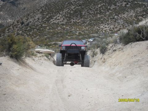 1990 Ford Bronco - A red off-road SUV navigates a dusty trail with other vehicles visible in the background.