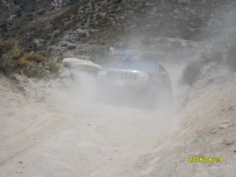 1990 Ford Bronco - Jeep Grand Cherokee navigates a dusty trail, with another vehicle in the distance.