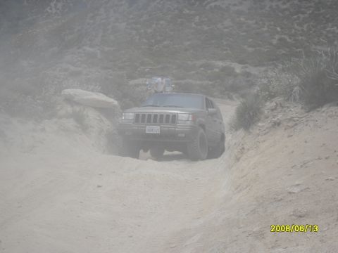 1990 Ford Bronco - A Jeep Grand Cherokee navigates a dusty off-road trail, with another vehicle following in the distance.
