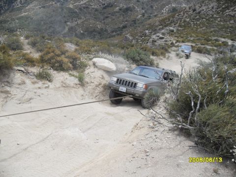 1990 Ford Bronco - A Jeep Grand Cherokee navigates a steep dirt trail, being assisted by a tow rope, with another vehicle visible further up the path.