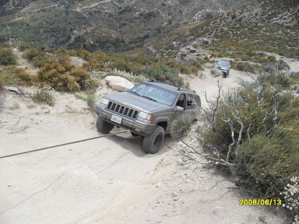 Gray Jeep Grand Cherokee on a steep dirt trail, being pulled by a rope. Another Jeep is visible further up the hill