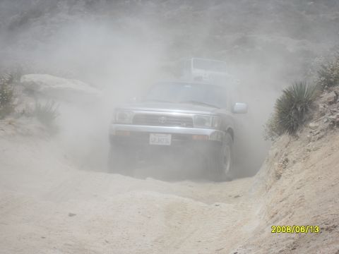 1990 Ford Bronco - A silver Toyota 4Runner kicks up dust on a dirt road, with another vehicle following closely behind.