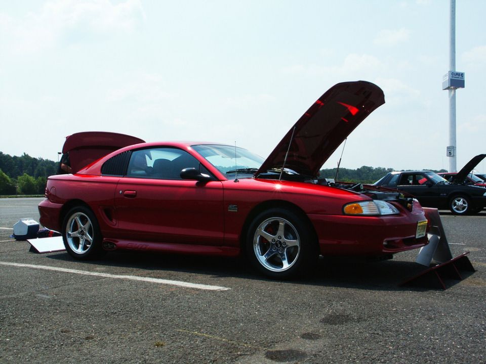 Red coupe with hoods open, side view, on asphalt. Another car is visible behind it