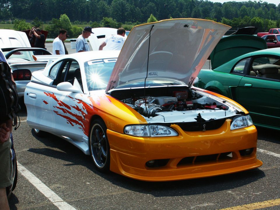 White and orange Mustang with flame decals, hood open showing engine. Another car in background