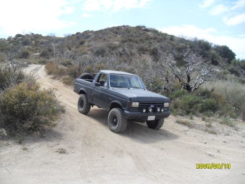 1990 Ford Bronco - This Ford Bronco is navigating a dusty trail with sparse vegetation and hills in the background.