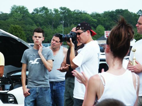 2001 Mercury Grand Marquis - A group of people are gathered around a car with its hood open at an outdoor event, with one person actively taking a photograph.