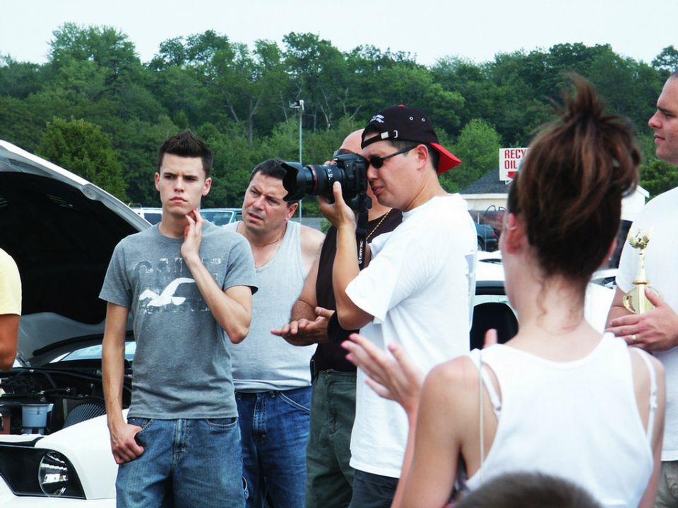 People gathered around a car with its hood up at an outdoor event, one person taking a photo