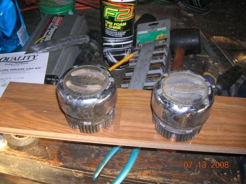 1985 Ford Bronco - Two chrome manual locking hubs are displayed on a wooden plank, with various tools and automotive products in the background.