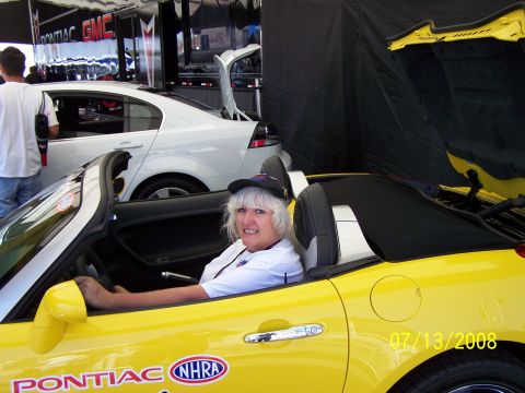 1972 Ford Ranchero - A woman sits in a yellow convertible adorned with NHRA and Pontiac decals, with a white sedan visible behind her.