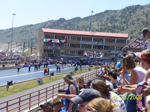 1972 Ford Ranchero - A drag racing event is underway at Bandimere Speedway, with spectators in the grandstands and a large building overlooking the track.