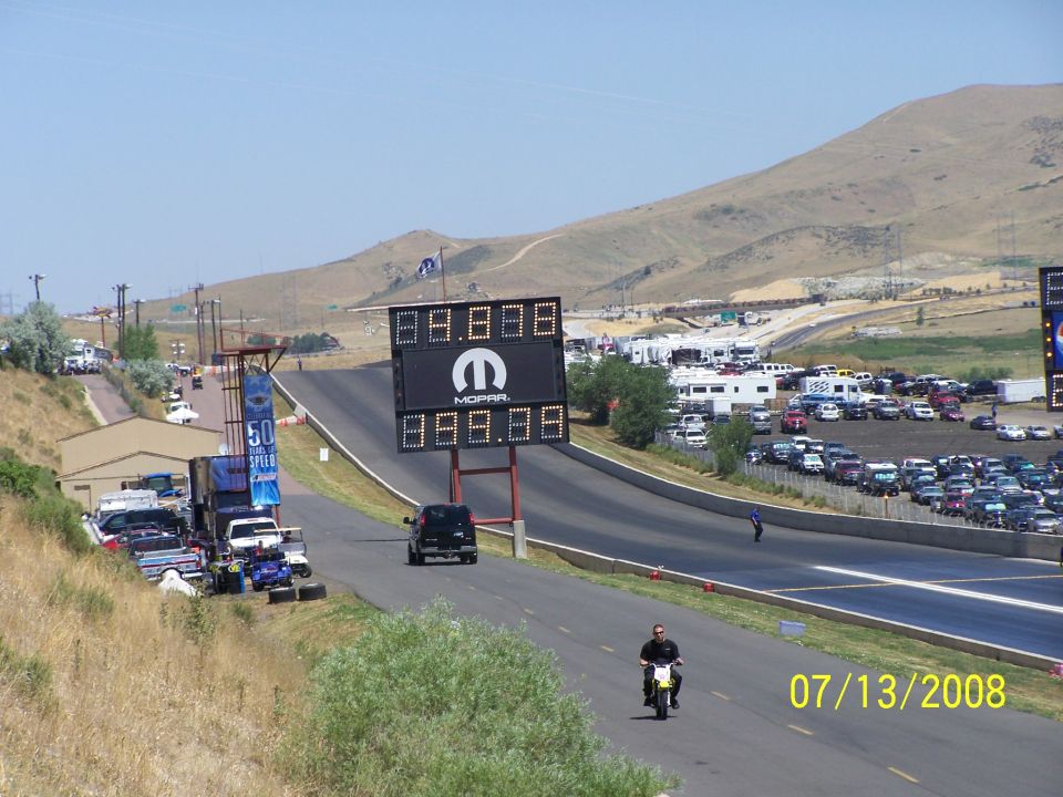 Drag strip with Mopar scoreboard, parked cars, RVs, and a motorcycle rider