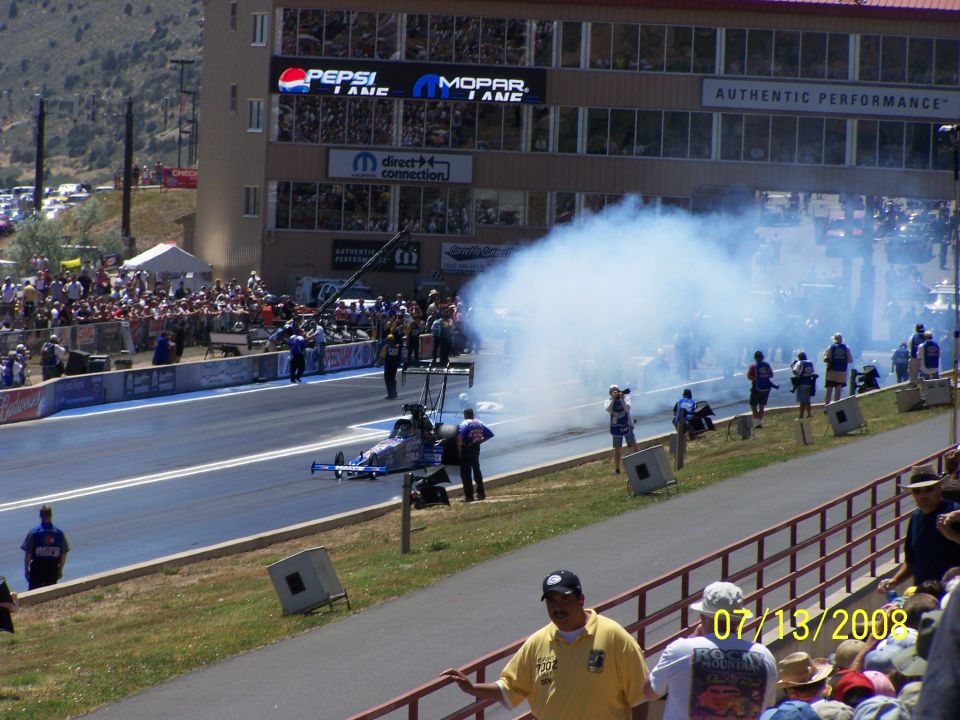 Drag race car on track with smoke, spectators in stands, and building with Pepsi and Mopar signs