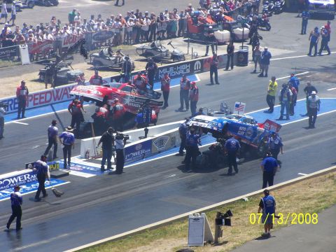 1972 Ford Ranchero - Two drag racing cars are attended to by crews on a track, with spectators watching from the stands.