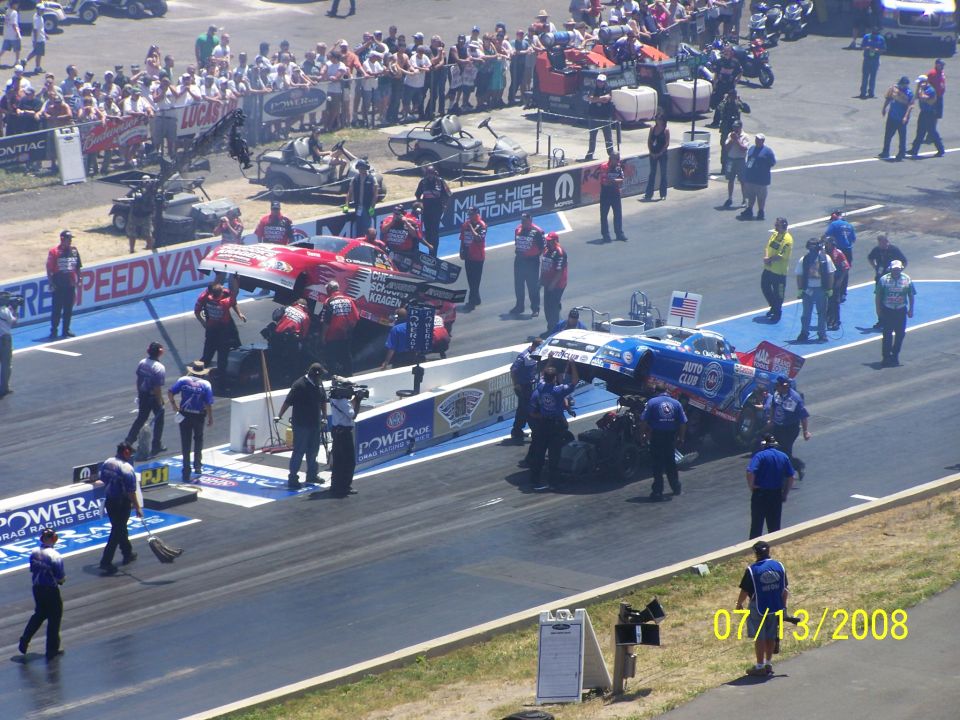 drag strip with two cars being worked on by crews, crowd in background