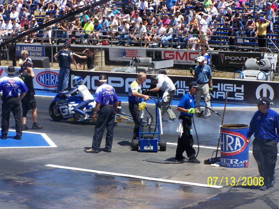 Drag racing motorcycle with rider on track, pit crew nearby, spectators in stands, date 07/13/2008