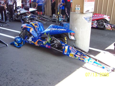 1972 Ford Ranchero - A custom drag motorcycle with extensive sponsor decals sits parked, with other vehicles and crew members visible in the background.