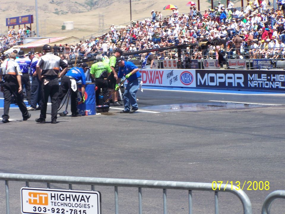 Drag strip with crew members and spectators in bleachers, with sponsor banners visible