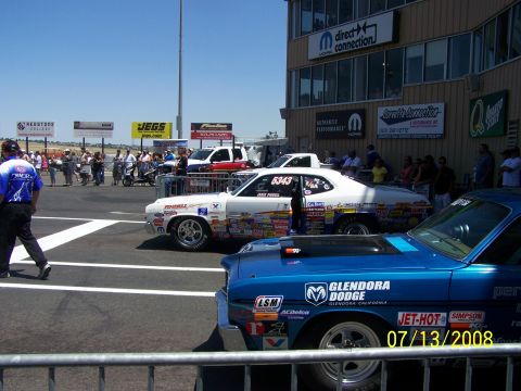 1972 Ford Ranchero - Two drag cars are lined up at a raceway, with a building and spectators in the background.