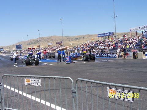 1972 Ford Ranchero - A drag racing event with multiple dragsters lined up on the track and a large crowd in the stands under a clear blue sky.