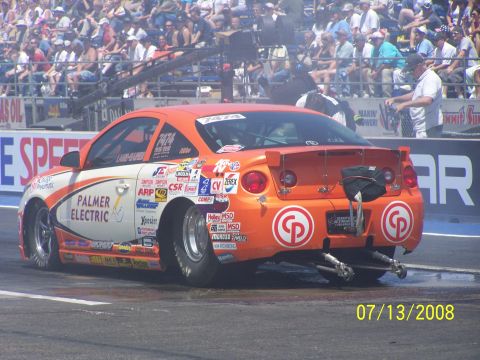 1972 Ford Ranchero - Orange drag car with numerous sponsor decals races down the track on a sunny day.