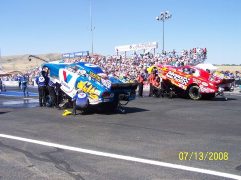1972 Ford Ranchero - Two drag racing cars are being serviced on the track during a competition, with a large crowd watching from the stands.