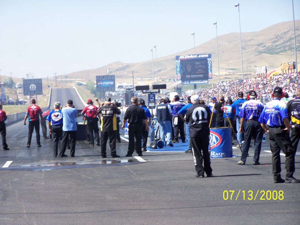 People in racing attire stand on a drag strip with a large crowd in the background. A digital scoreboard is visible