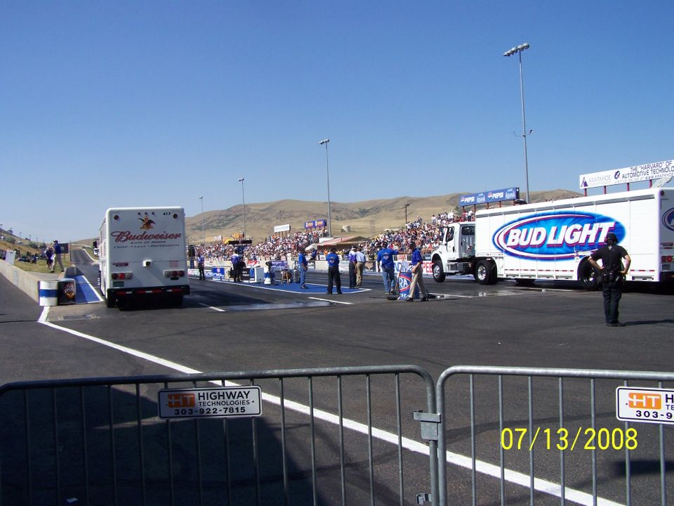 Two large trucks, one with Budweiser and one with Bud Light logos, parked at a drag strip with spectators in the background
