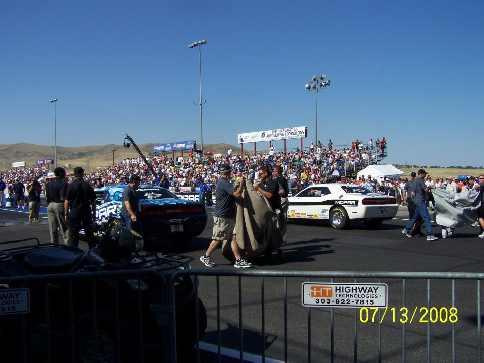 Two drag cars on a track with a crowd watching. One car is black with blue graphics, the other is white with black stripes and Mopar logos