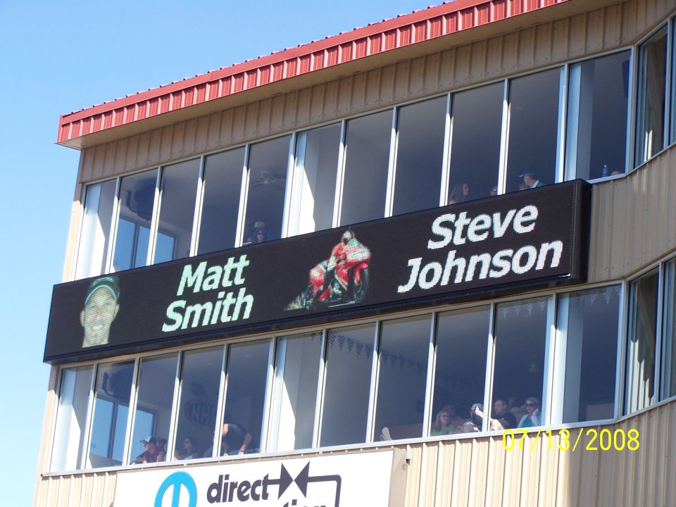 Digital billboard shows Matt Smith and Steve Johnson with a motorcycle image between them. Building has many windows with people inside