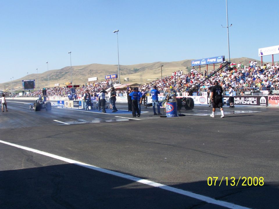 Drag strip with two dragsters at the starting line, spectators in the stands, and mountains in the background. Date stamp reads 07/13/2008