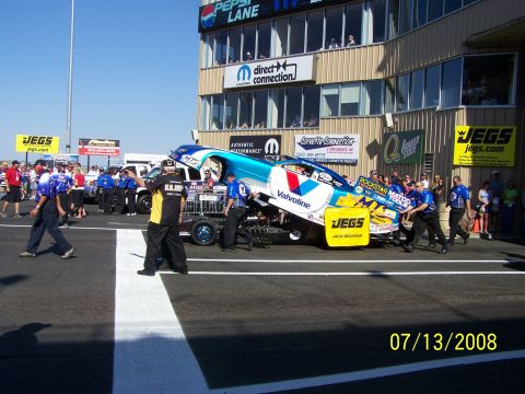 1972 Ford Ranchero - A drag racing car is being pushed by a crew on a track in front of a building with various sponsor signs.
