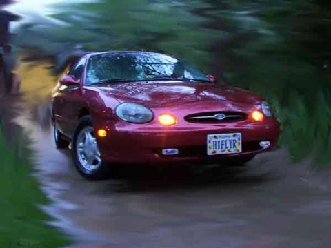1998 Ford Taurus - A red sedan drives down a dirt road, kicking up dust.