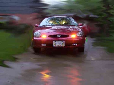 1998 Ford Taurus - A red 1998 Ford Taurus is seen from the front, with its headlights illuminated on a wet surface.