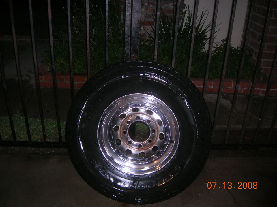 Chrome alloy wheel with 8 lug pattern and black tire, wet, in front of a black metal fence